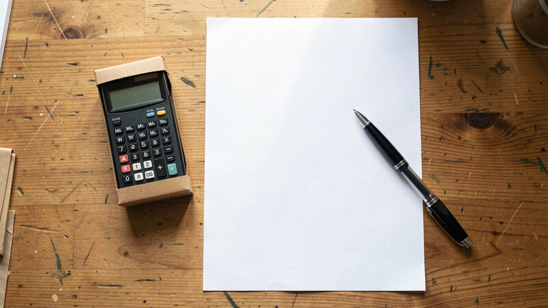 A photograph of several different banknotes and coins spread on a wooden desk beside a small parcel and a calculator, top-down view